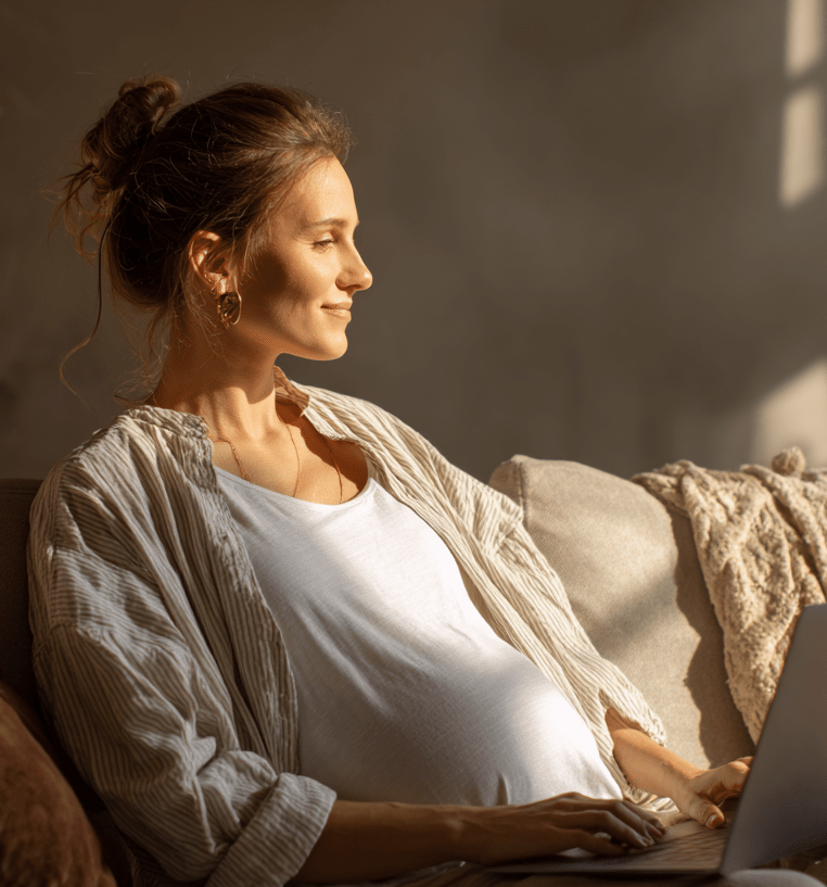 Schwangere Frau sitzt entspannt auf dem Sofa mit Laptop auf dem Schoß und genießt die Sonne.